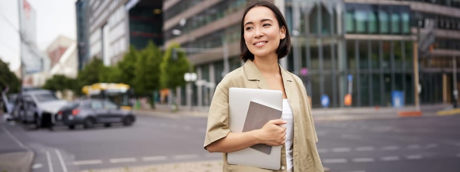 Portrait of beautiful asian woman standing on street with laptop and notebook, going to university or work, commute route.