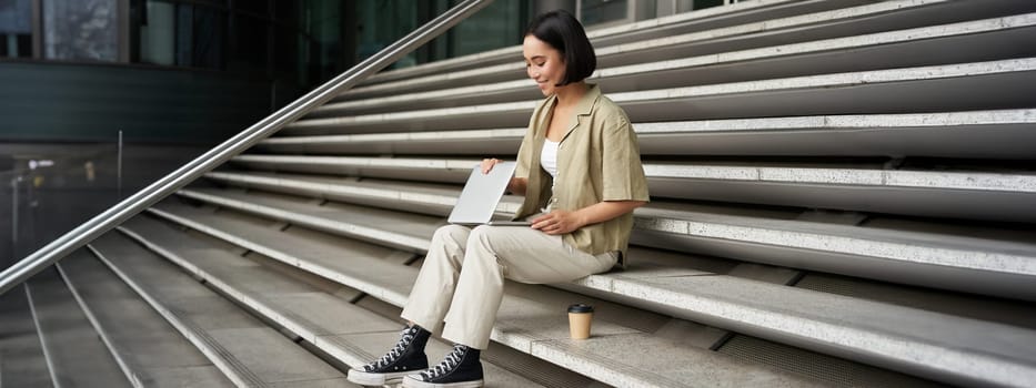 Young smiling asian girl with laptop, sits on street of city with coffee. Young woman does homework on computer while sitting on street stairs, digital nomad works remote.