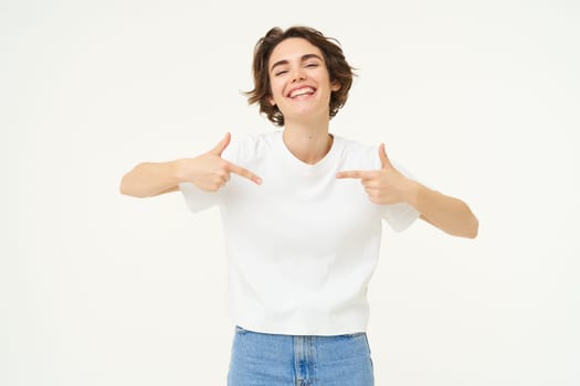 Portrait of smiling, confident girl, pointing at herself with pleased face, standing over white studio background. Copy space