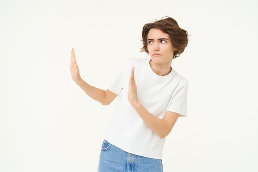 Portrait of woman avoiding someone, shows block hands gesture and refusing from something bad, standing over white background.