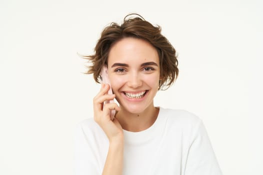 Image of cute brunette woman answer phone call, talking on smartphone, holding mobile near eat and smiling , standing over white background.