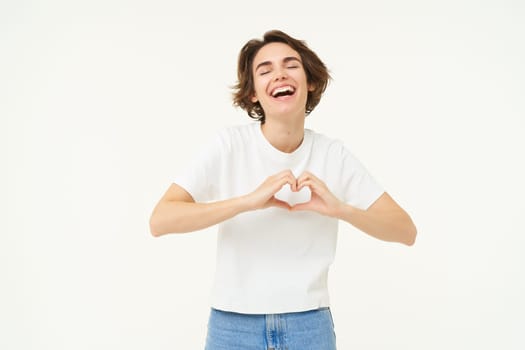 Portrait of young woman shows heart, love and care gesture, express sympathy, like something or someone, smiling happily, standing over white background.