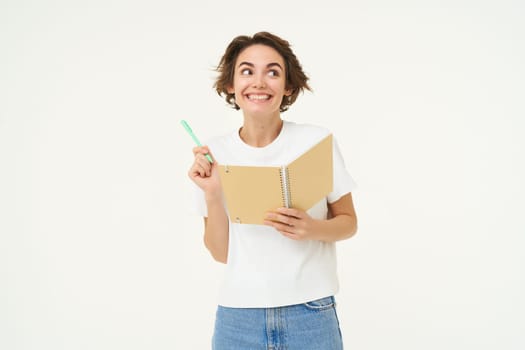 Portrait of smiling, beautiful and creative woman, holding pen and notebook, writing in her planner, has a diary, posing over white studio background.