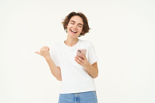 Portrait of smiling, carefree woman, holding smartphone, pointing finger at copy space, advertisement, using mobile phone and recommending store, standing over white background.