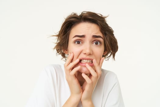 Close up of scared brunette woman, shaking from fear, looking concerned and frightened, standing over white studio background.