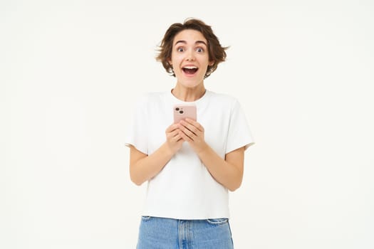Portrait of brunette girl with shocked face, reading something amazing on mobile phone, standing with smartphone over white background.