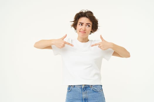 Portrait of unsure brunette girl, pointing at herself and looking uncertain, looking away, standing over white studio background. Copy space