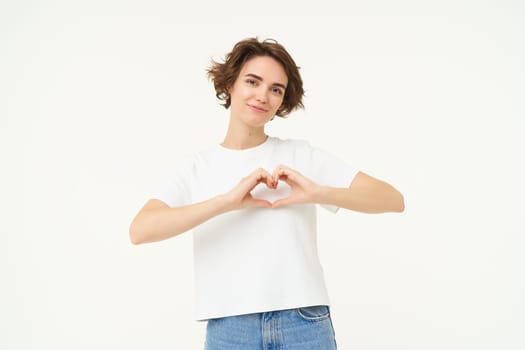 Portrait of brunette girl shows heart sign, love gesture, express care and warm feelings, stands over white background.