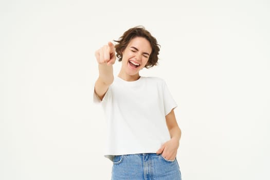 Portrait of excited, smiling young woman, pointing finger at camera, inviting you, congratulating, standing over white studio background.