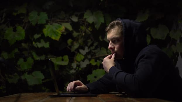 A young man at a table in a gazebo works on a laptop during late at night. A man in a black hoodie with a laptop against a background of green grapes. 4k