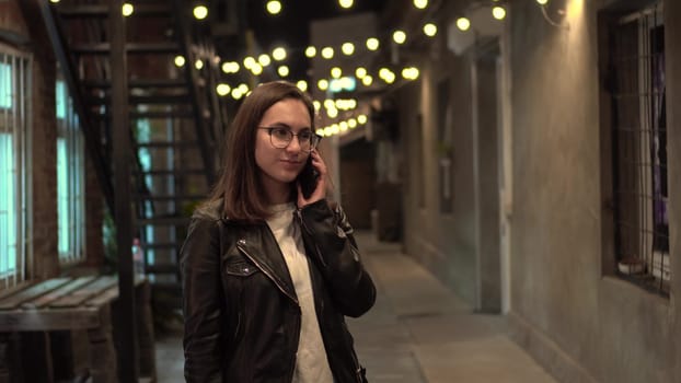 A young woman talks on the phone on a narrow street in the evening. A girl with glasses and a leather jacket on a cozy street. 4k