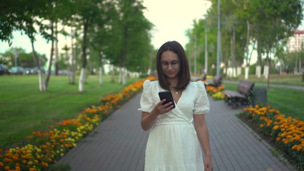 A young woman walks along a flower alley with a phone in her hands. A girl in a white dress looks at her phone and walks among the flowers. 4k