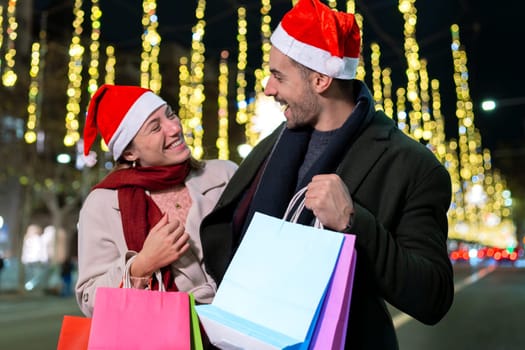Smiling couple wearing Santa hats, standing under festive Christmas lights. They look at each other warmly, creating a joyful holiday atmosphere. One holds a phone, capturing memories.