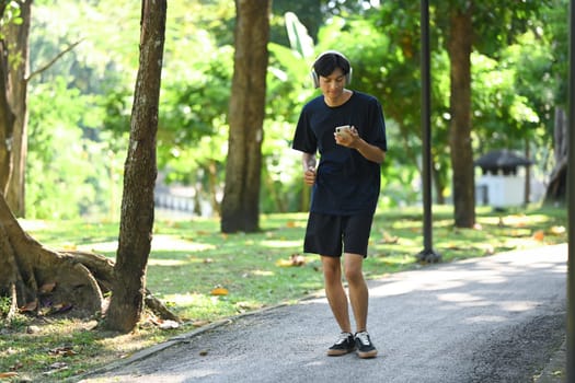 Young man holding mobile phone jogging along a paved path in a park on a sunny day.