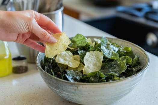 Woman grabbing a delicious spinach chip from a bowl, enjoying a healthy snack in a modern kitchen