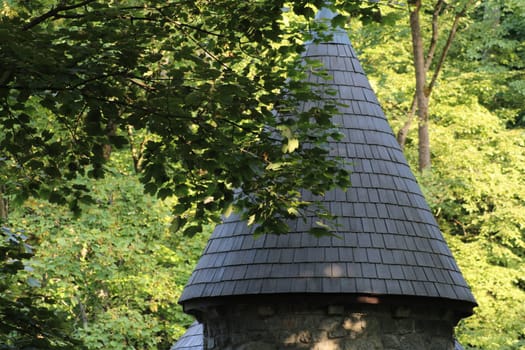 The stone tower features a distinctive conical roof and is nestled among vibrant green trees, creating a tranquil atmosphere in this forested area on a sunny day.
