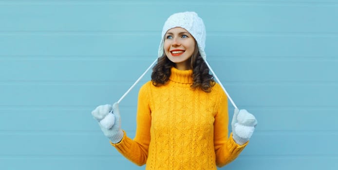 Winter christmas portrait of happy young woman smiling in yellow sweater, white hat looking away standing on blue background