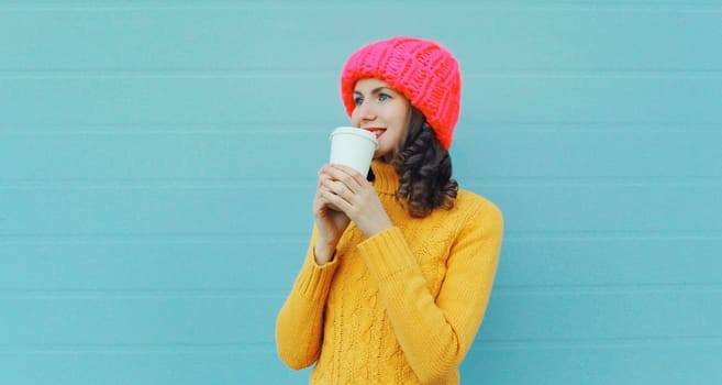 Winter portrait of happy smiling young woman in pink hat, yellow sweater with cup of coffee drink looking away standing on blue background