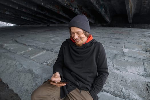 A cheerful individual sits on a concrete surface beneath a bridge, engrossed in their smartphone, smiling and enjoying the simple pleasure of connecting with others.