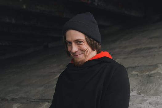 A man with long hair and a beard sits quietly under a bridge, lost in thought, wearing a beanie and hoodie. The urban setting evokes calm despite the bustling city above.