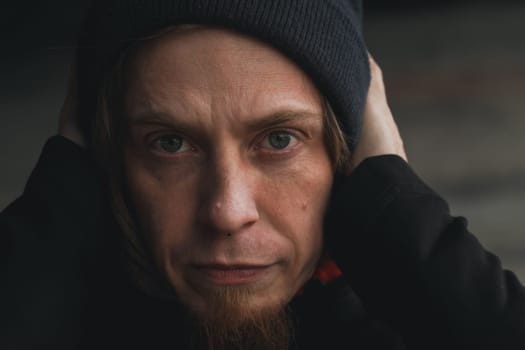 In a moody urban atmosphere, a young man with long hair in a beanie covers his ears, emulating a moment of introspection and vulnerability amidst the chaos.