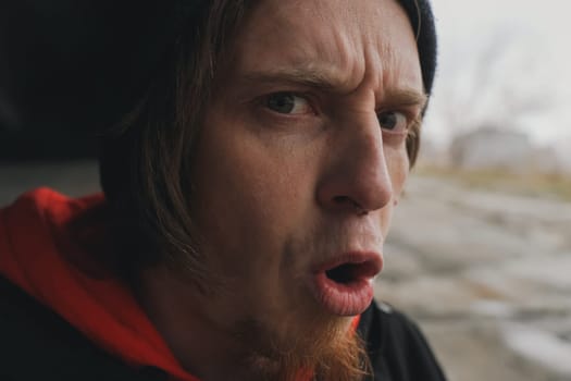 Expressing astonishment, a man with a beard wearing a hooded sweatshirt reacts to something unexpected in a deserted location on an overcast day.