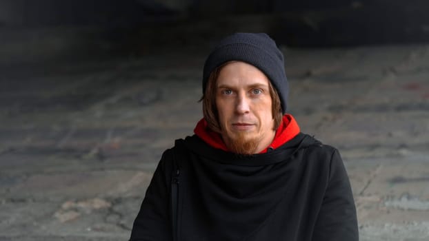 In a dimly lit urban space, a man gazes thoughtfully while wearing a red hoodie and a beanie.
