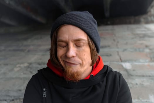 A man with long hair and a beard sits quietly under a bridge, lost in thought, wearing a beanie and hoodie. The urban setting evokes calm despite the bustling city above.