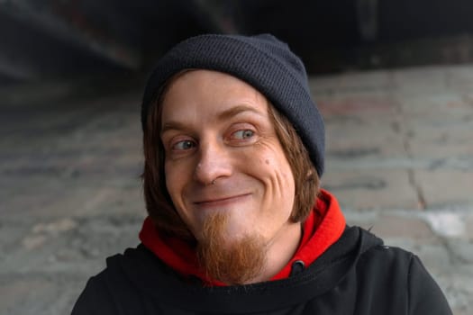 A man with long hair and a beard sits quietly under a bridge, lost in thought, wearing a beanie and hoodie. The urban setting evokes calm despite the bustling city above.