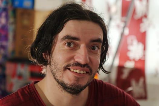A cheerful man with shoulder-length dark hair beams with joy in a colorful indoor environment filled with vivid decorations, creating a warm atmosphere.