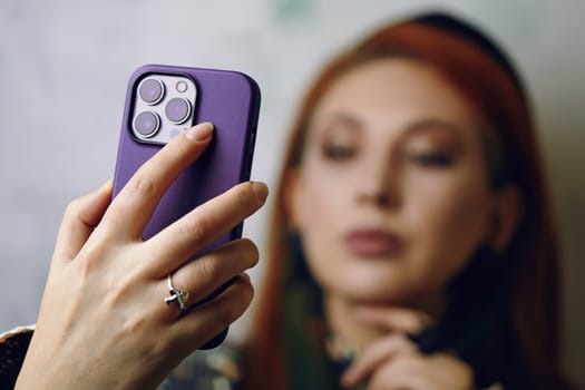 Red hair woman takes selfie on iPhone 13 pro. Close-up of female's hand with mushrooms ring holding smartphone in violet case. Gadget with triple-lens camera. Bishkek, Kyrgyzstan - January 24, 2022