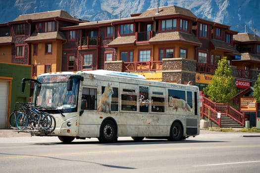 A tourist bus with bicycles attached to the front rides along a road in the town of Canmore in the mountains of Banff National Park in Canada. Canmore, Canada - 08.03.2023