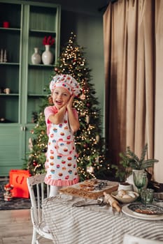 .a girl child in the kitchen with a Christmas tree is preparing or cooking ginger cookies for Christmas or New year