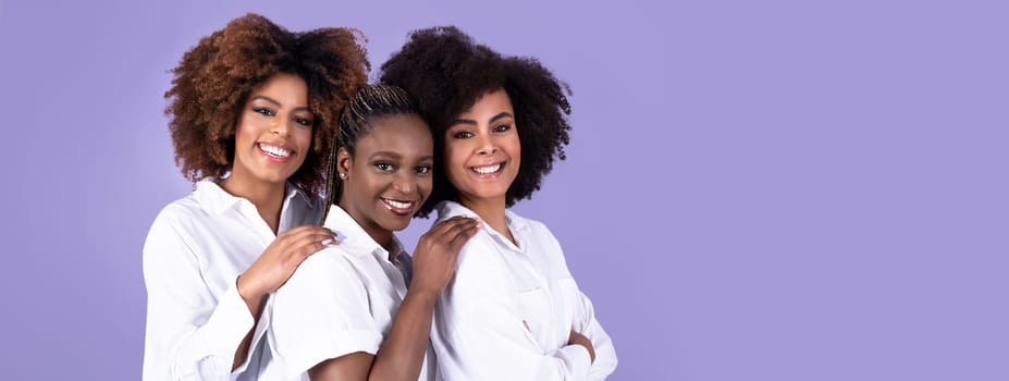 Beauty and togetherness. Three happy united black young women smiling to camera, posing and hugging over purple studio background, wearing white shirts, portrait shot of ladies trio, copy space