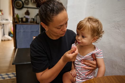 A mother lovingly feeds her toddler in a cozy kitchen. The child is seated on her lap, wearing a striped outfit, while she gently holds the spoon. The scene captures a moment of nurturing and care.