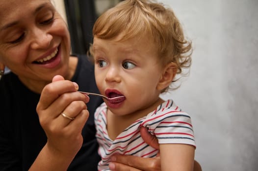 A mother lovingly feeds her toddler son, who looks curious while tasting food from a spoon. The cozy indoor setting adds warmth to the moment, highlighting maternal care and the bonding experience.