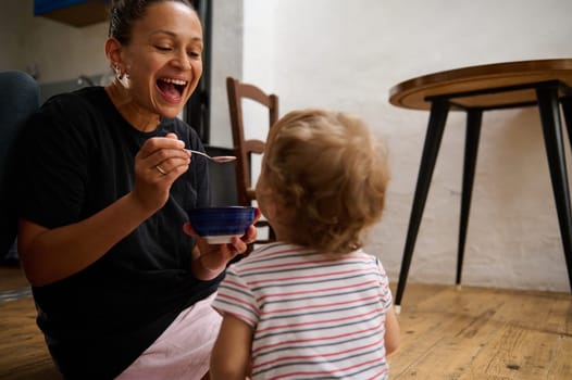 A joyful mother engages with her toddler during mealtime on the kitchen floor, sharing moments of laughter and bonding. The scene captures love, warmth, and family togetherness.