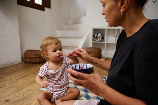 Mother feeds her toddler daughter with care and tenderness, sitting in a warm home setting. The child is enjoying a meal, showcasing a loving bond. The scene radiates comfort and warmth.