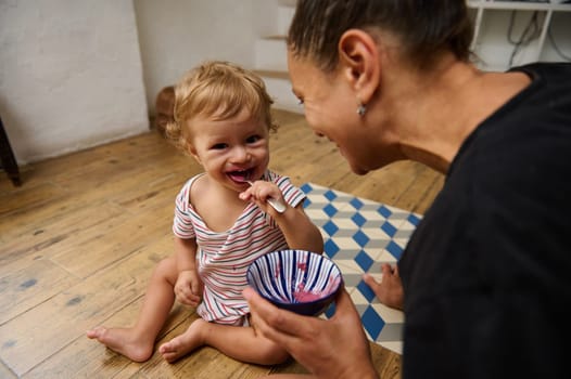 A delightful scene of a parent joyfully feeding their toddler in a cozy home setting. The child's bright smile and the colorful bowl of food create a warm, family-focused atmosphere.