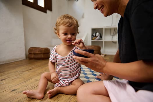 A mother feeds her adorable toddler with a smile in a cozy home interior. The child enjoys a meal in a warm, loving atmosphere, showcasing family bonding and joyful parenting moments.
