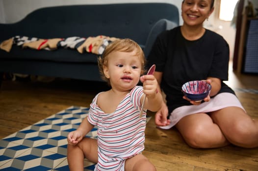 A joyful scene of a toddler exploring painting while sitting on the floor, accompanied by a smiling caregiver. The room exudes warmth and creativity.