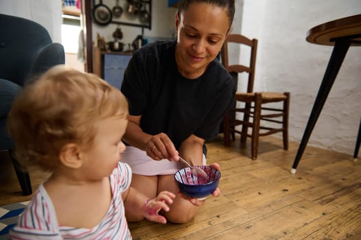A tender moment of a mother feeding her young child in a cozy kitchen environment. The child is dressed casually and enjoys a playful interaction. The room is warmly lit, reflecting a homely ambiance.