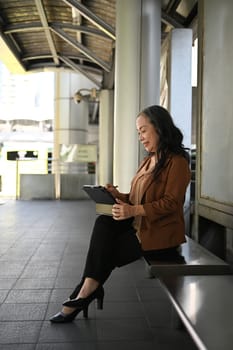 Smiling mature businesswoman working using laptop at a bus station.
