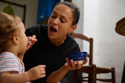 A mother lovingly feeds her baby with a spoon in a cozy kitchen setting, showcasing warmth and care. The child's curiosity and the mother's tenderness highlight a nurturing bond.