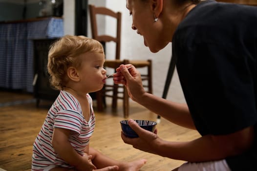A tender moment as a mother lovingly feeds her toddler sitting on the wooden floor of a cozy kitchen. The toddler looks curious and content. The room exudes warmth and intimacy.