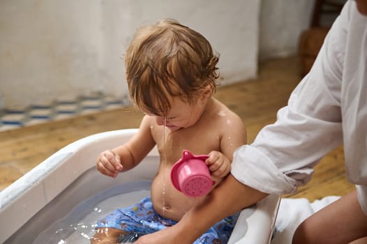 A cute toddler plays with a pink watering can during bath time. The child is in a small tub, surrounded by warm indoor lighting, highlighting the joyful and innocent moment.