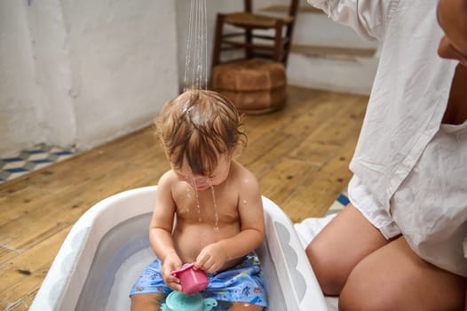 A cute toddler sits in a small bathtub, playfully enjoying water streaming down their head, creating a joyful and carefree atmosphere in a cozy room.