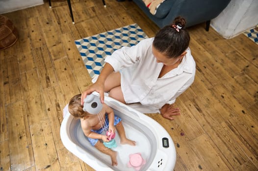 A mother lovingly bathes her toddler in a small tub placed in the living room. The cozy setting features a warm wooden floor and a nearby sofa, emphasizing a caring and nurturing environment.