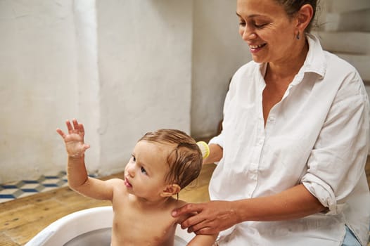 A happy mother lovingly bathes her baby in a warm and cozy home environment. The baby raises a hand, exploring the surroundings, while the mother smiles gently, creating a tender family moment.