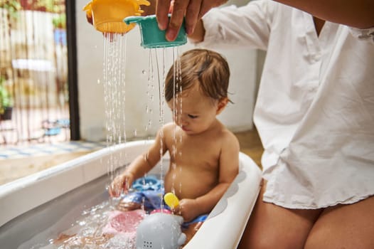 A toddler enjoys playful bath time with a parent, pouring water from colorful cups. The scene captures childhood curiosity and family bonding in a cozy home setting.